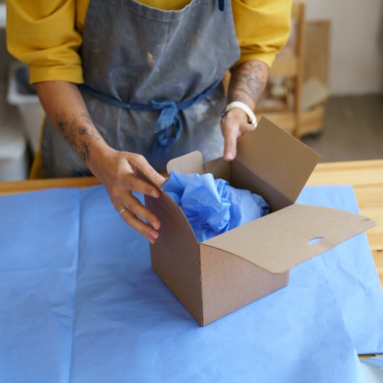 Deliver parcel: girl potter artist in apron packing ceramics in box with paper for safe shipping